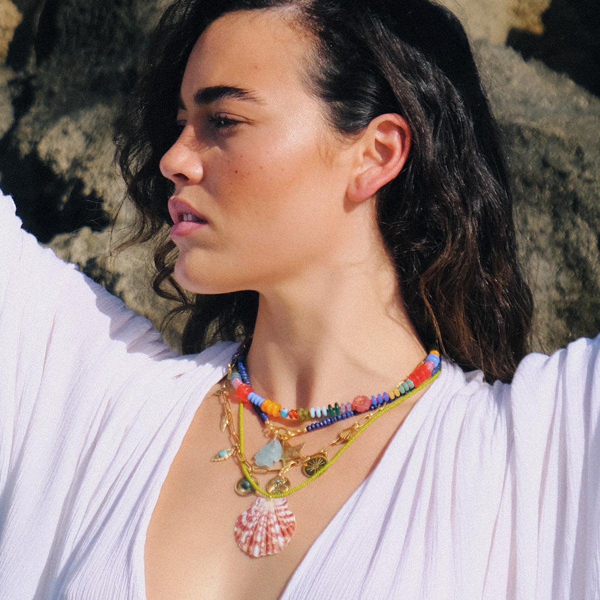 A woman with wavy brown hair wears a white top and layered necklaces, including the Rainbow Candy Necklace—a handmade piece with colorful glass beads and a large shell pendant. She stands outdoors in sunlight, rocks behind her, looking aside with raised arms.