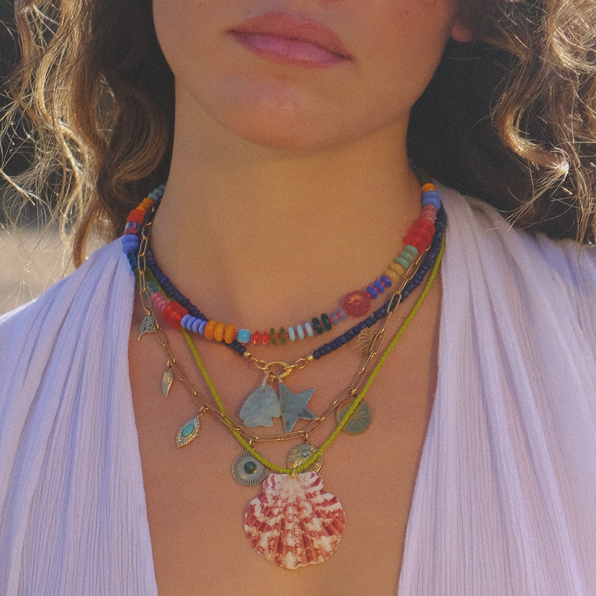 A woman in a white top wears layered necklaces, including the Rainbow Candy Necklace, a handmade beaded piece with glass beads, colorful charms, a blue fish pendant, and a large reddish-brown seashell. Her face is partially visible.