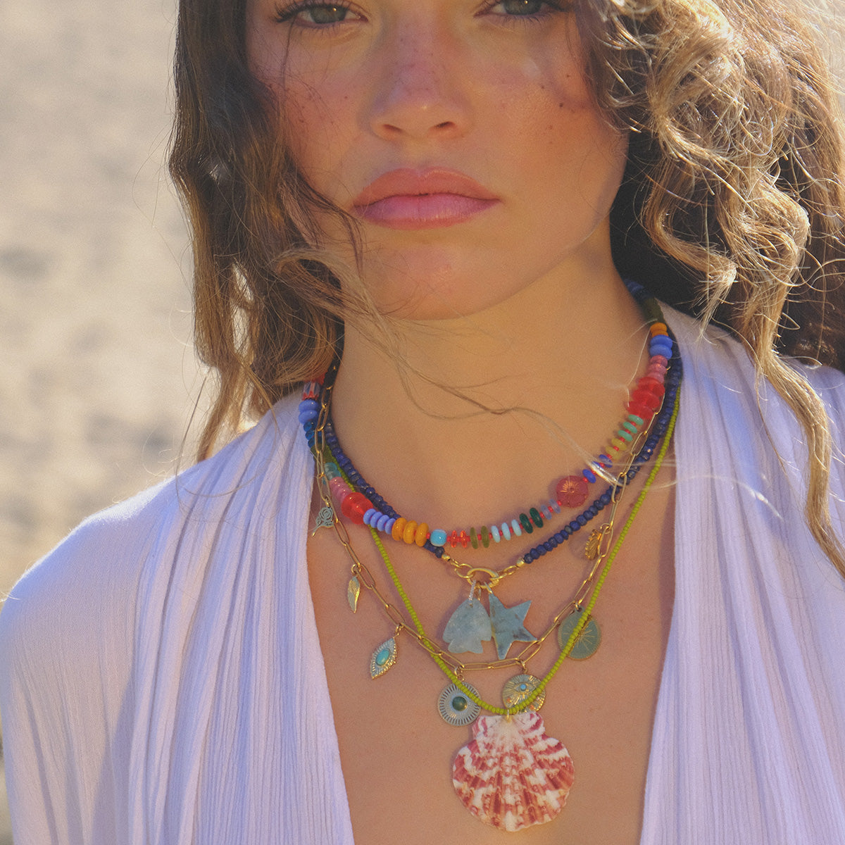 A woman with wavy hair and a white top wears the Rainbow Candy Necklace—layered, colorful handmade beads featuring shells, charms, pendants, and glass beads—against a sunlit sandy background.