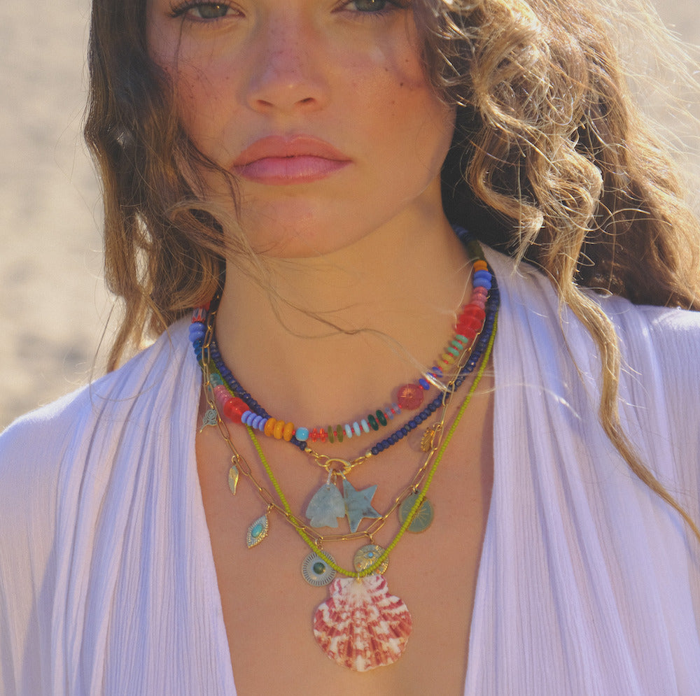 A woman with wavy hair wears the Beaded Gemstone Charm Holder Necklace, featuring layered beads and sea-inspired charms like a shell and starfish, over a white v-neck top against a sunlit sandy background.