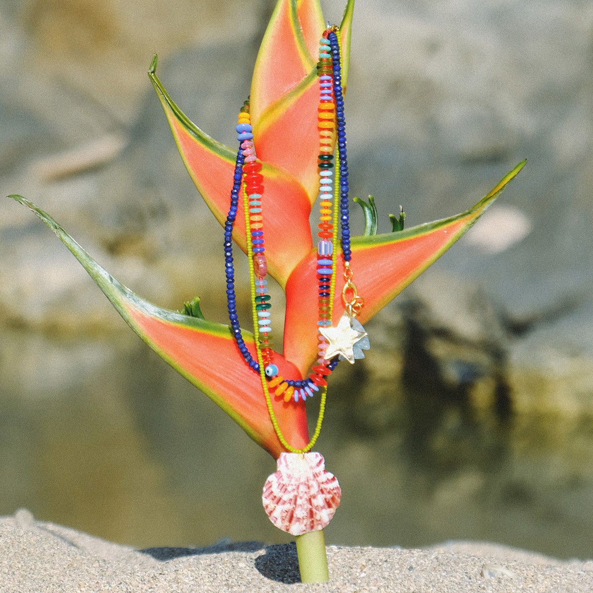 The Rainbow Candy Necklace, a colorful handmade piece with star and shell charms, features glass beads and a pink seashell charm. Shown draped on a vibrant orange tropical flower, its bright colors pop against a sandy, blurred background.