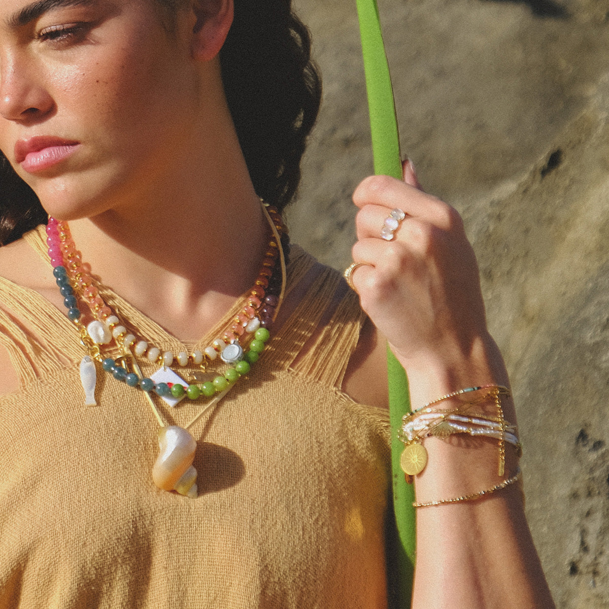 Wearing layered Miyuki bead necklaces and the Single Serpent Beaded Bracelet, a person with curly hair in a tan fringed top holds a green leaf, standing in sunlight against rocky terrain.
