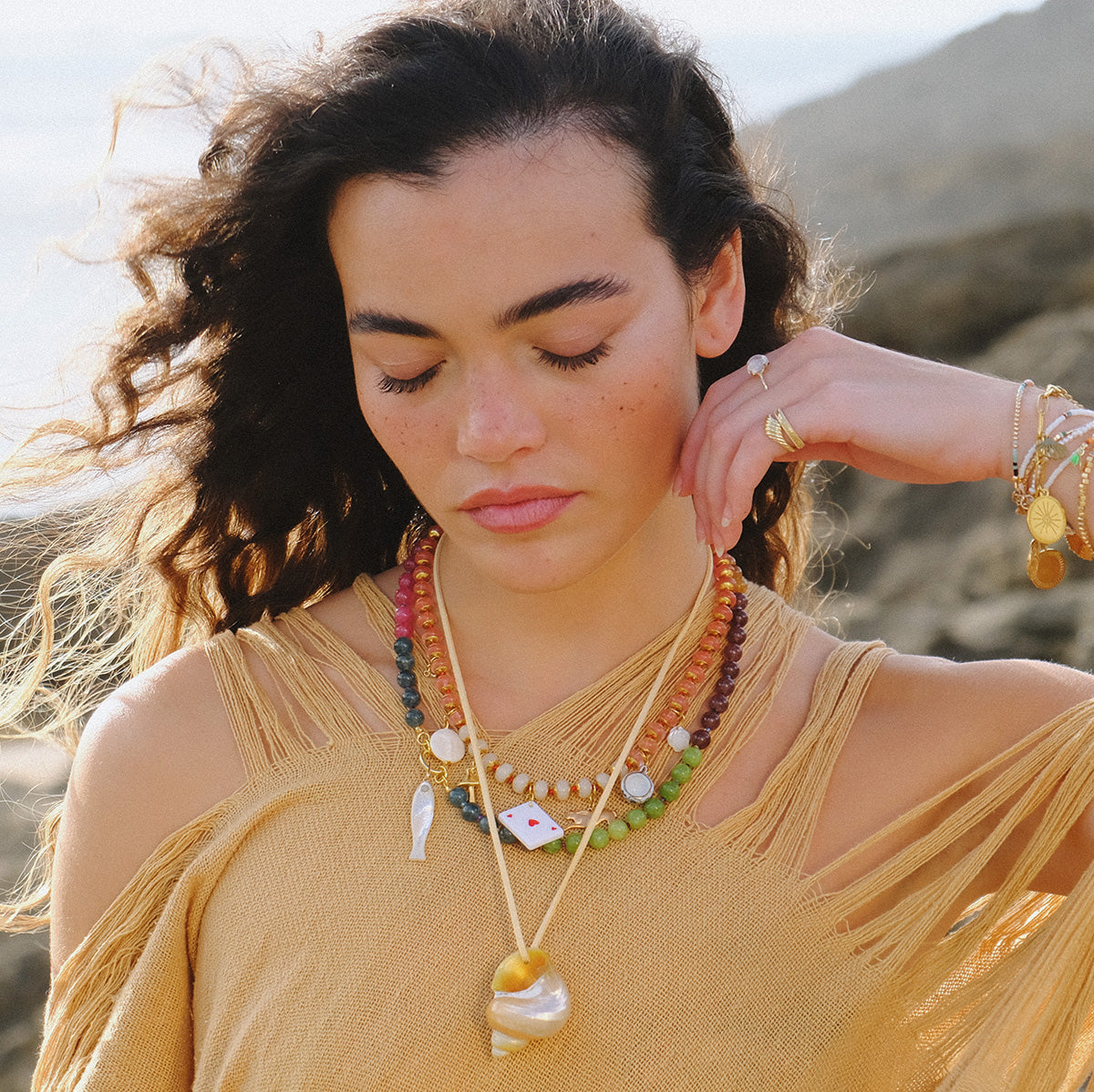 A woman with wavy brown hair and freckles wears the Wonderland Necklace, paired with shell jewelry and a tan fringed top. Outdoors, she stands with her eyes closed, touching her hair as rocks and sky fill the background.