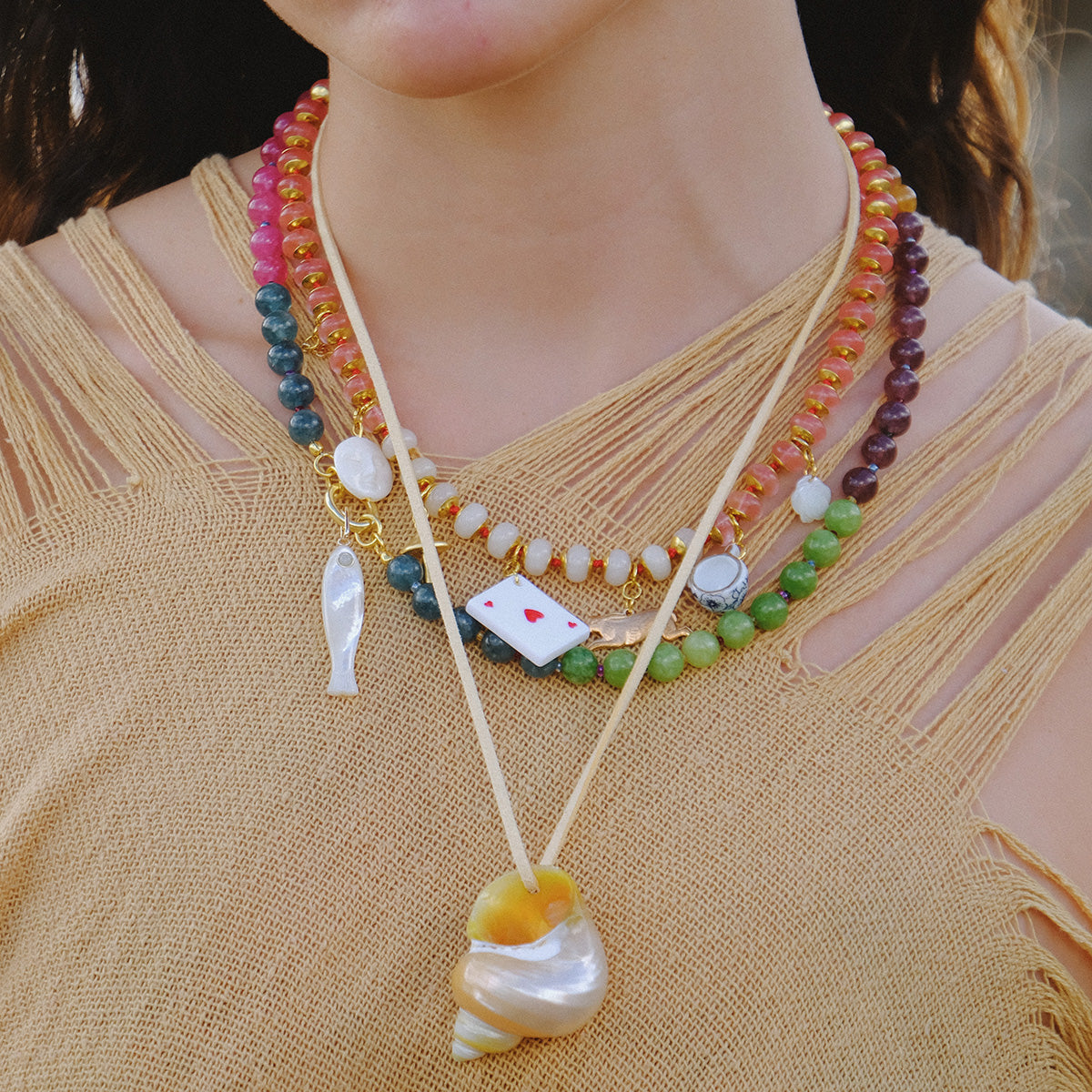A person wearing a tan, fringed top and several colorful beaded necklaces with unique pendants, including a fish, a Wonderland Necklace styled like a playing card, and a large shell.