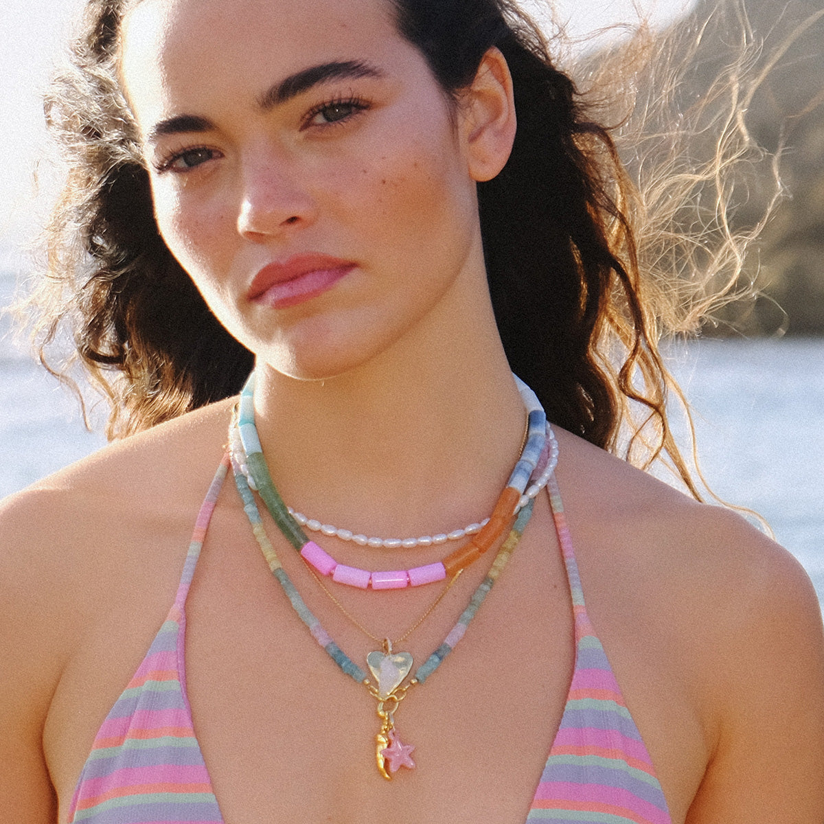 A young woman with wavy hair wears layered beaded necklaces, featuring the 14k Pink Rhodochrosite and Diamond Sea Star Charm, paired with a pastel striped halter top. She stands outdoors by water and rocks, her calm face adorned with natural makeup.