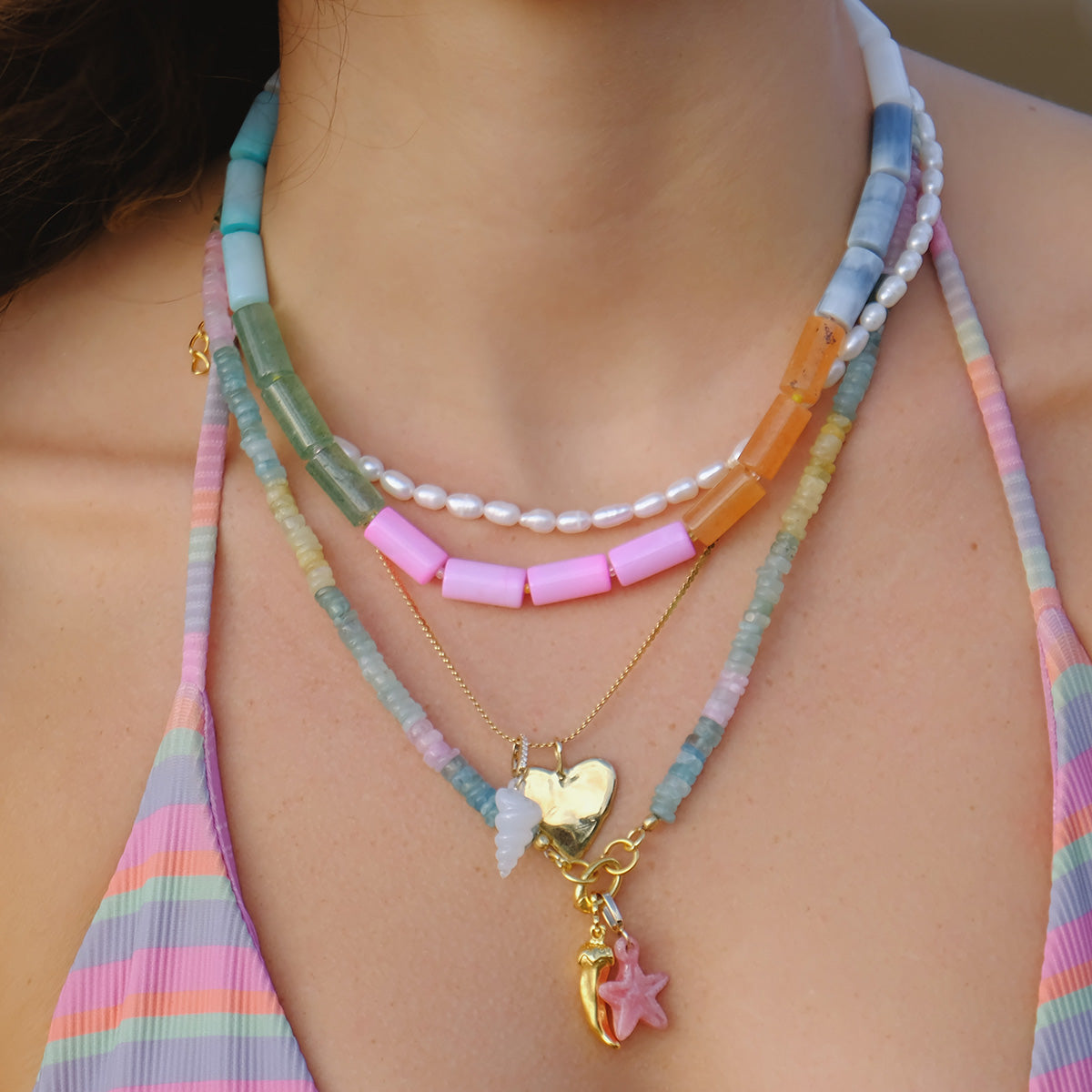 A close-up of a person wearing layered necklaces, including the 14k Pink Rhodochrosite and Diamond Sea Star Charm, with colorful beads, pearls, and gold pendants over a pastel striped top.