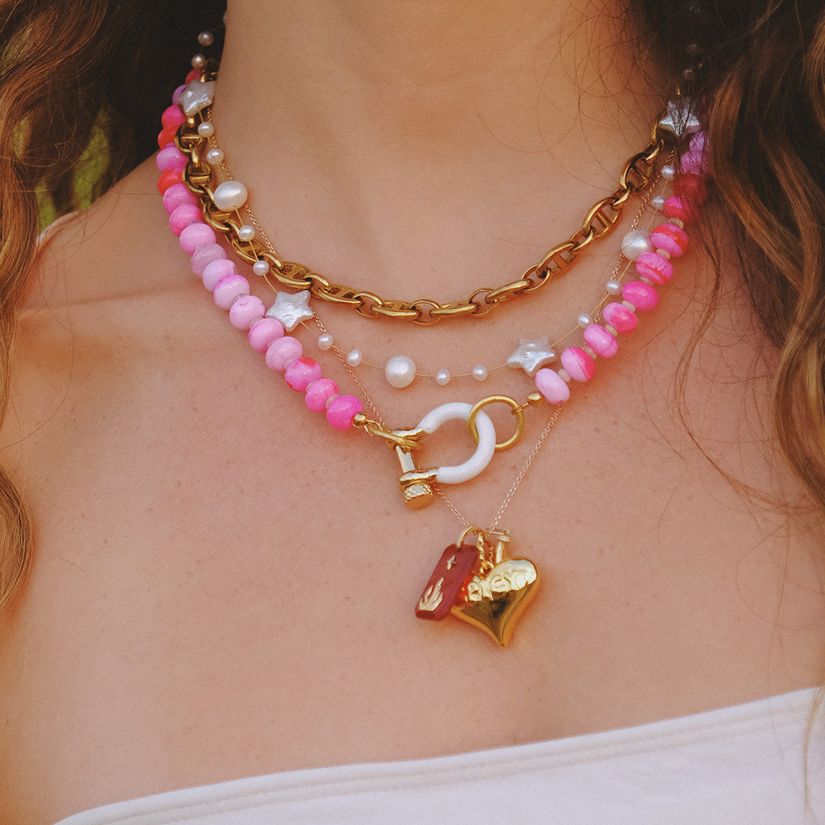 A woman in a white strapless top wears layered necklaces: a gold-plated chain, the pink-beaded Suki Necklace, one with natural pearls and star beads, and two gold pendants—a heart and a rectangular charm.