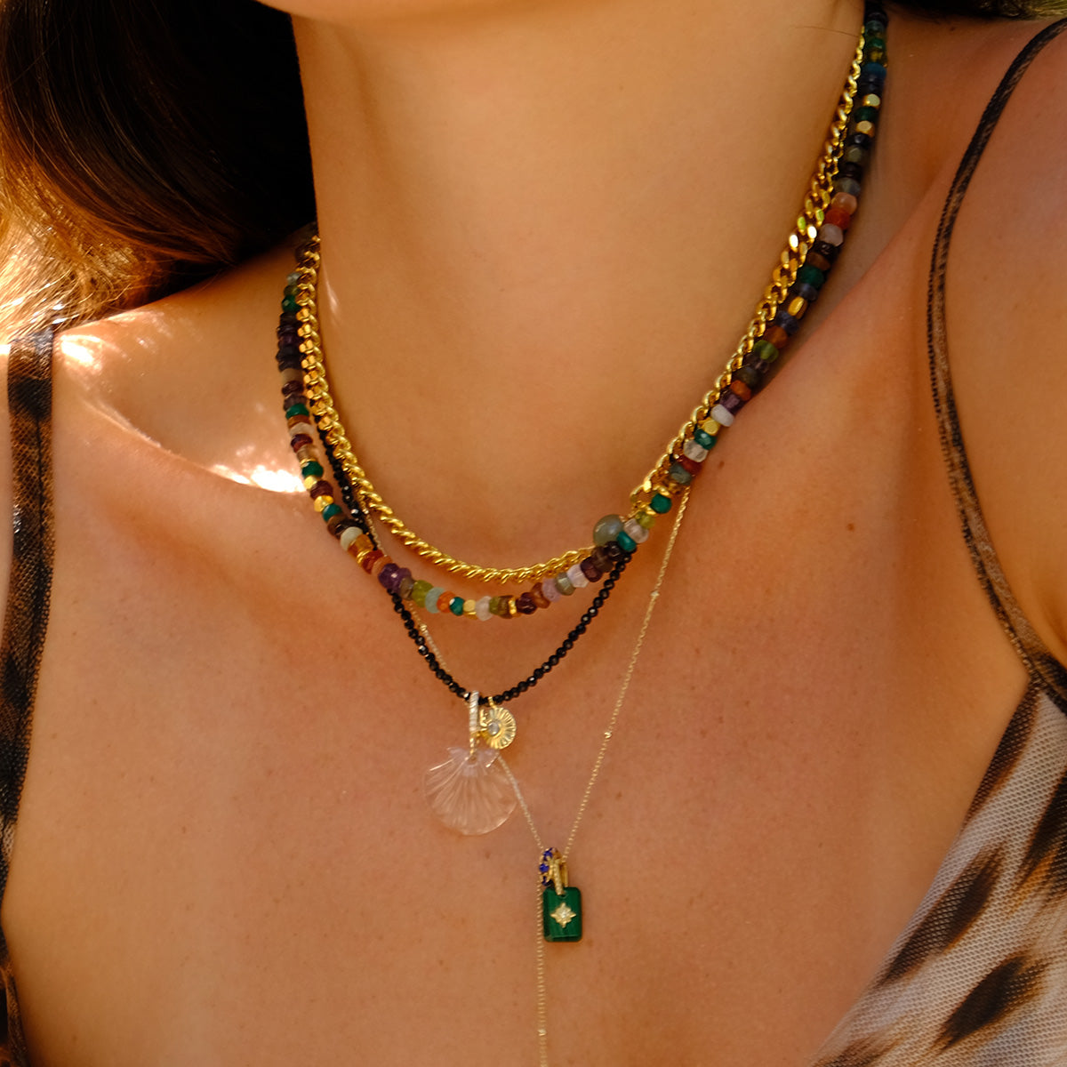 A woman wearing the Solid Gold Beaded Lariat Necklace, styled with gold chains, multicolored beaded strands, a crystal pendant, and a green charm; sunlight highlights her skin and patterned clothing.