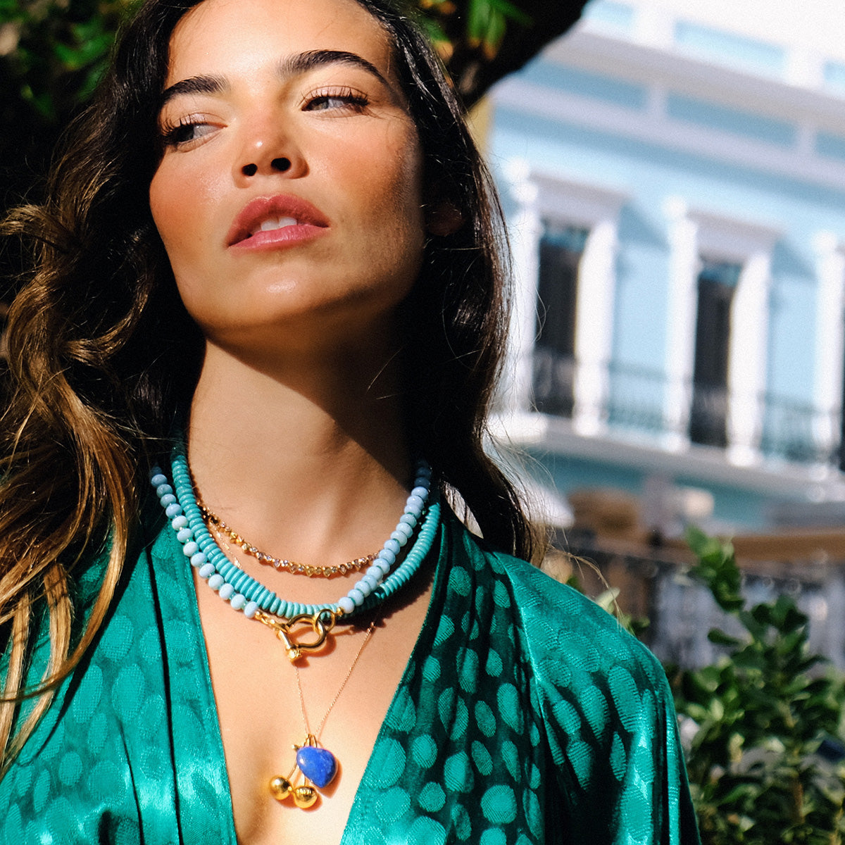 A woman in a green patterned top wears the Teardrop Tennis Necklace, a layered 14k gold piece adorned with cubic zirconia, standing outdoors in sunlight with a blurred building behind her.