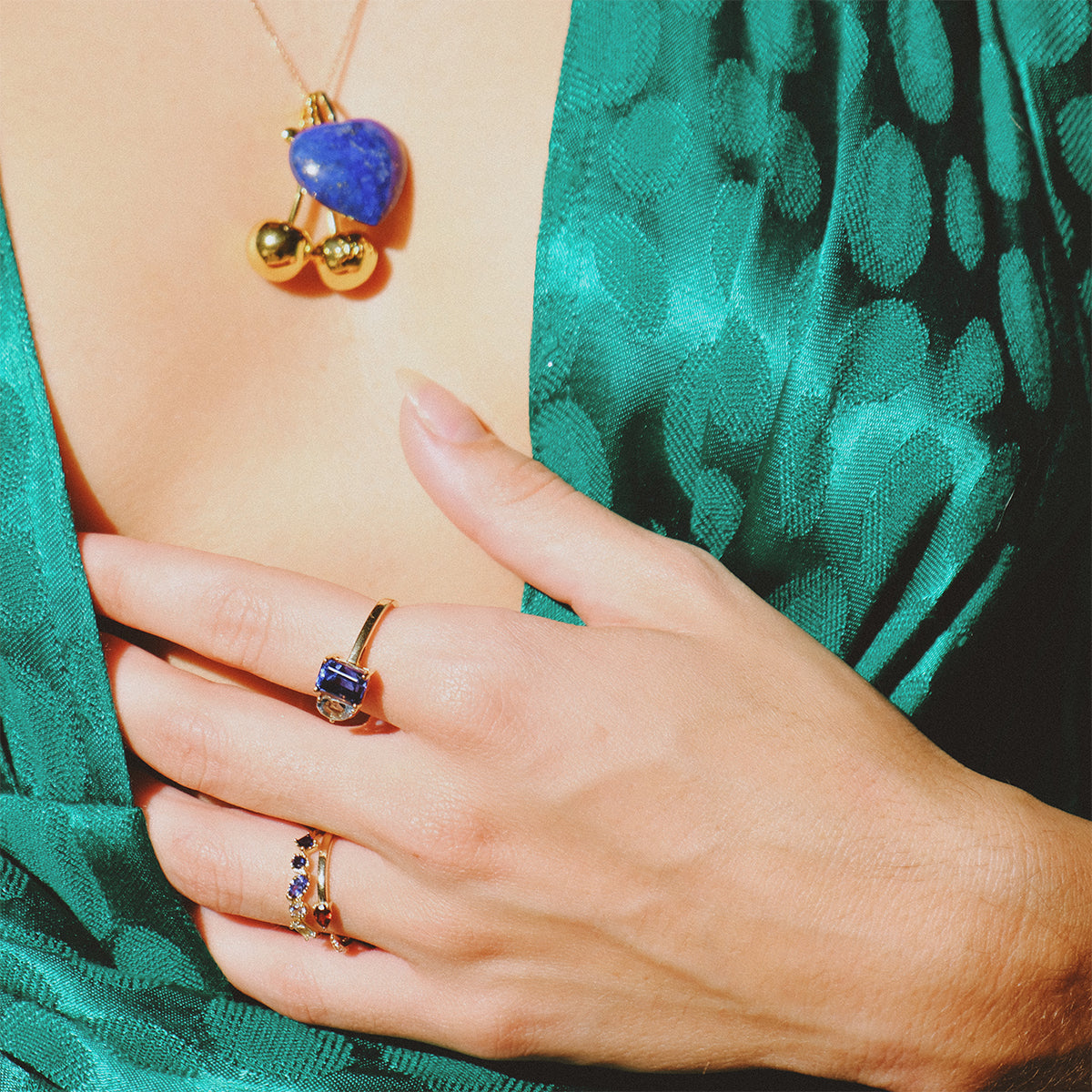 A person wearing a green satin dress, 14k gold plated Shades of You Ombre Birthstone Rings with blue and purple stones, and a gold necklace featuring a blue heart and gold cherries pendant, hand resting on their chest.