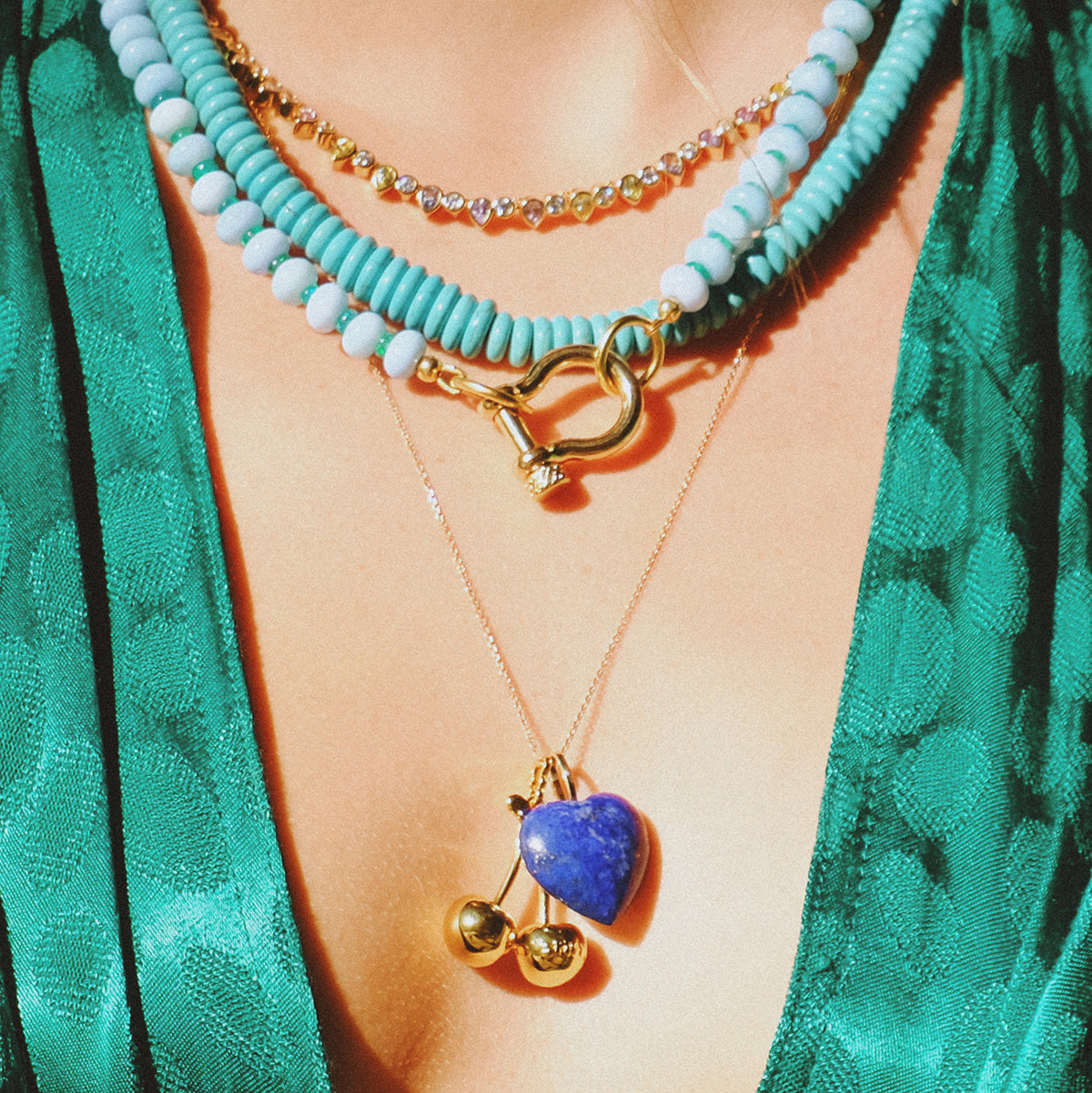 A person wearing a green textured garment with layered necklaces: one featuring turquoise beads, a 14k gold chain with cubic zirconia gems, and the Teardrop Tennis Necklace adorned with a blue heart pendant and gold sphere charms.