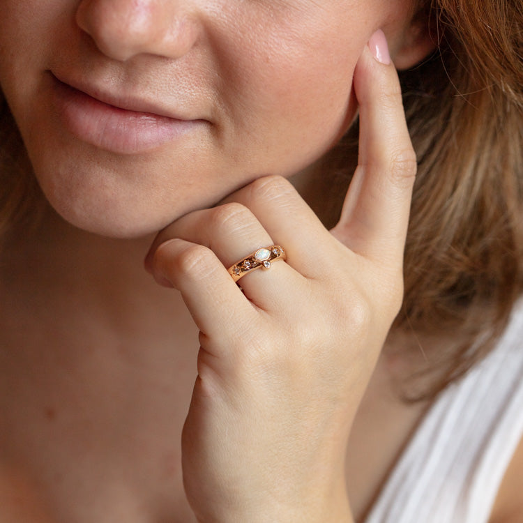 A woman with light skin and light pink nails gently touches her face, showcasing the Athena Opal Ring Sample Size 8, a 14k yellow gold plated ring with a round synthetic opal and a 4 mm band, while wearing a white top.