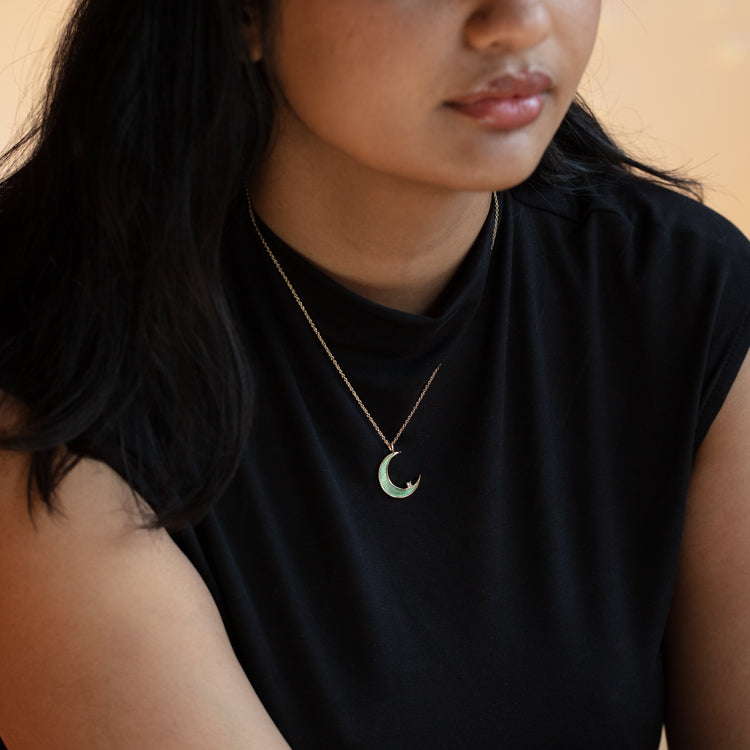 A woman with long dark hair wears a black top and showcases the La Lune Teal Green Necklace, featuring a crescent moon pendant. The softly lit image highlights her necklace and upper torso.
