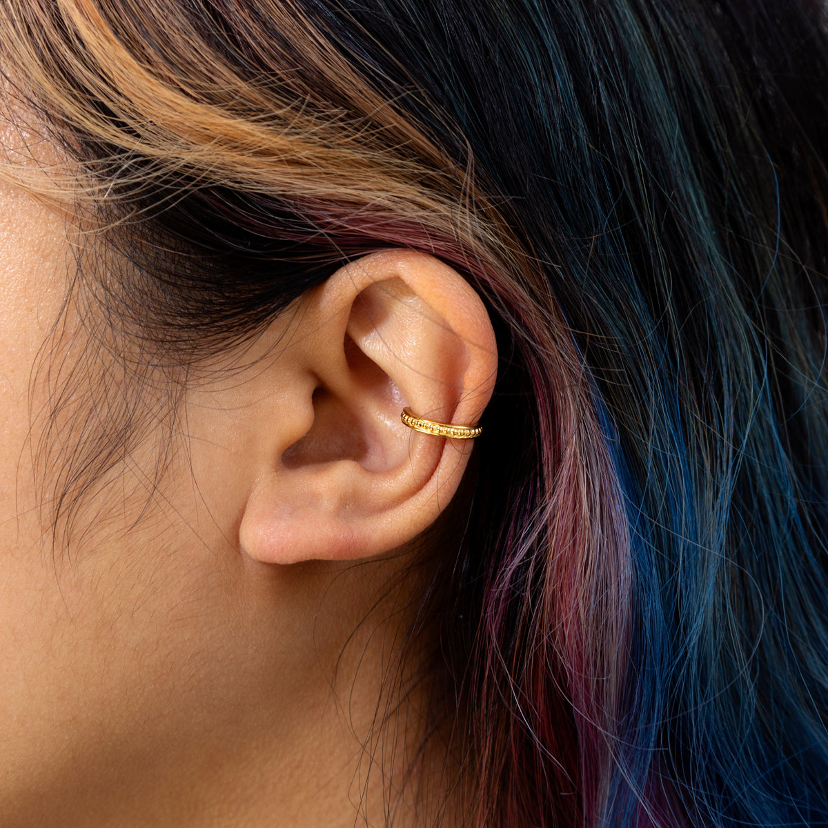 Close-up of a person’s ear wearing the Beaded Ear Cuff Sample, a 10.5 mm helix cuff with a textured 14k yellow gold plated finish over a silver base. The wearer has dark hair with blonde and blue highlights, background blurred.