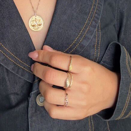 A person in a dark jacket holds the lapel, displaying gold rings—including the Shades of You Ombre Birthstone Ring—and a 14k gold plated pendant necklace featuring a scales of justice design.