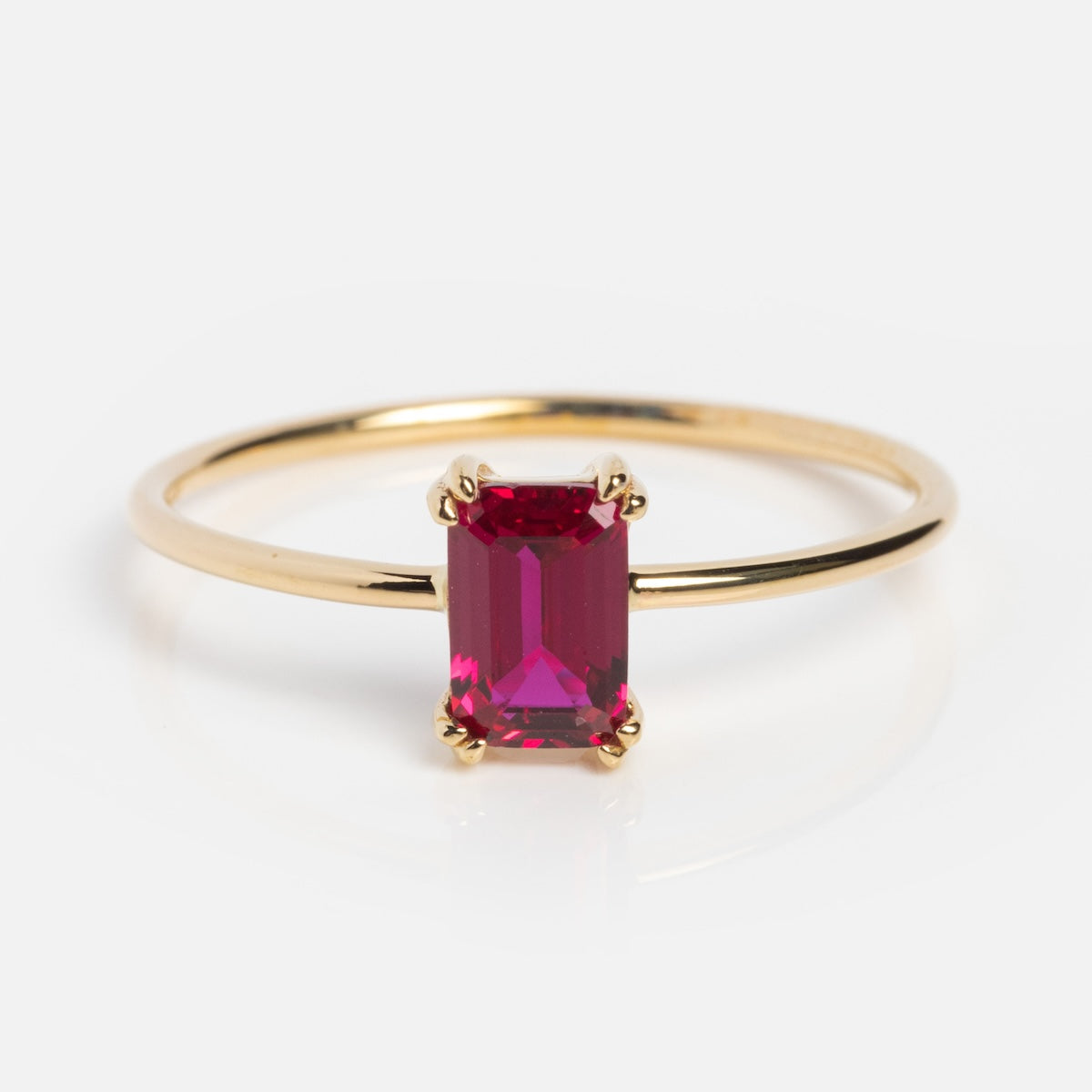 The Solid Gold Essential Baguette Birthstone Ring features a thin solid gold band with a vertically set rectangular emerald-cut red gemstone in a four-prong setting, shown on a white background.