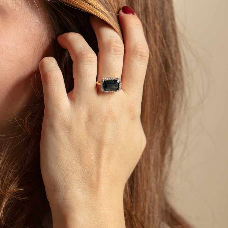 A person with brown hair touches their ear, displaying the 14k Solid Gold Diamond East West Baguette Ring featuring a large rectangular gemstone and sparkling diamonds on their finger.