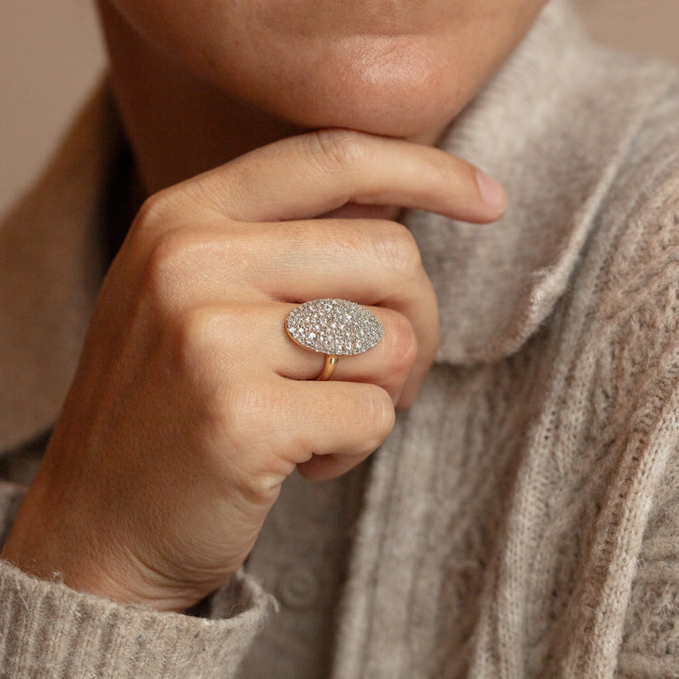 A person in a cozy light gray sweater rests their chin on their hand, showcasing the 14k Solid Gold Bella Diamond Navette Ring—an elegant, modern piece featuring a large oval diamond.
