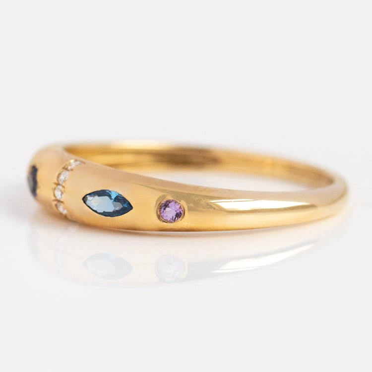 The Solid Gold Pizza Acrostic Ring features marquise-cut blue stones, a round purple gem, and tiny clear stones set in a curved gold band, inspired by classic acrostic jewelry and shown against a white background.