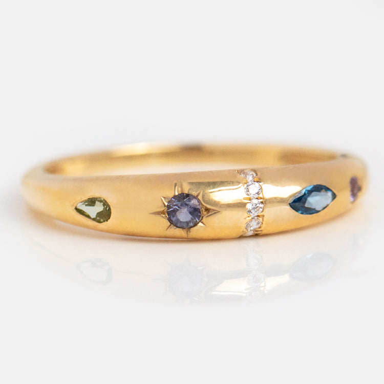 The Solid Gold Pizza Acrostic Ring showcases a central blue stone, vertical white diamonds, and an array of blue, green, and purple gemstones set around the band for a vibrant statement jewelry piece.
