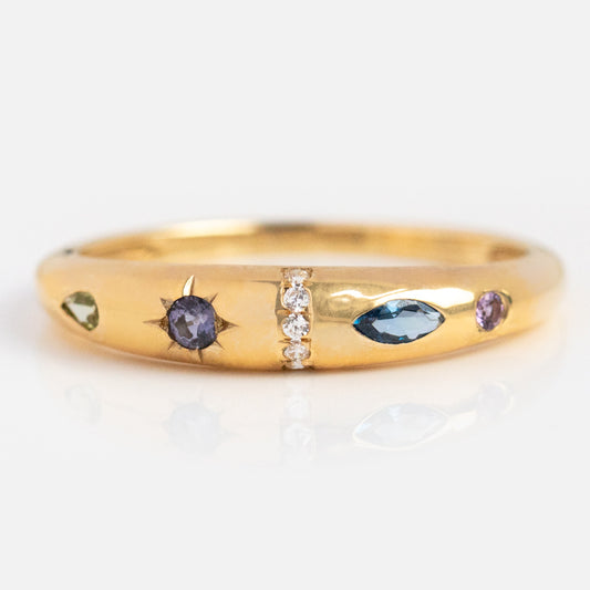 The Solid Gold Pizza Acrostic Ring features a round purple stone in a starburst setting, a marquise blue stone, rows of clear gems, and vibrant accents—capturing the playful elegance of modern acrostic jewelry.