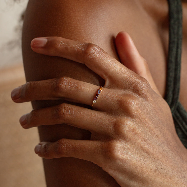 A close-up shows a hand with medium brown skin resting on a shoulder, wearing the Solid Gold Magic Acrostic Ring Sample Size 7—crafted in thin 9k solid gold and set with small amethyst gemstones. The person wears a dark green sleeveless top.