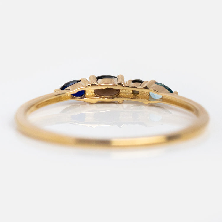 The Solid Gold Boss Acrostic Ring (sample size 7) is a thin 9k gold band with a decorative top set with small colored gemstones, including blue topaz, shown in side view on a white background with detailed settings and delicate band visible.