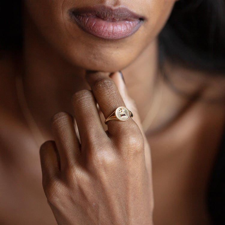 A close-up of a woman’s hand touching her chin, wearing the Solid Gold Quhier Que Demain Signet Ring. Her lips and part of her face are visible, complemented by a delicate gold necklace.