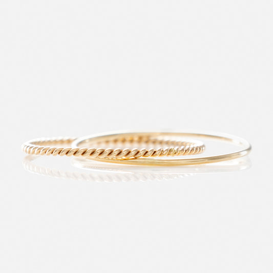 Two solid gold bands—a twisted rope and a smooth design—overlap on a white backdrop, casting soft reflections. The Solid Gold Crossover Ring Set is a Local Eclectic exclusive that radiates timeless elegance.