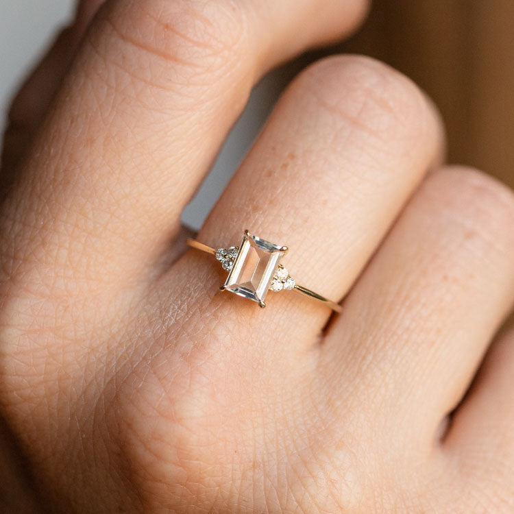 A hand models the Solid Gold Essential White Topaz Ring Sample, featuring a rectangular white topaz center stone flanked by two small round white diamonds, set in yellow gold against a neutral background.
