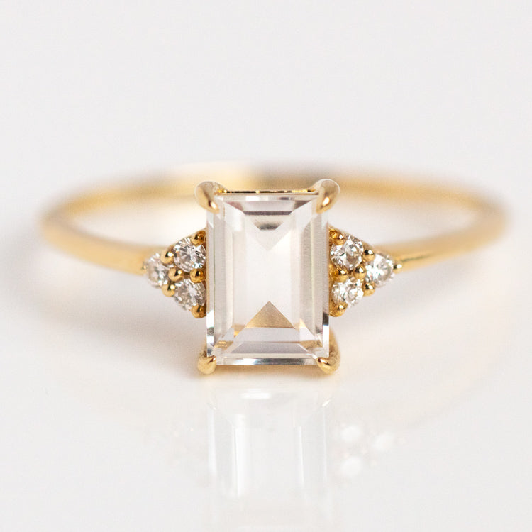 The Solid Gold Essential White Topaz Ring Sample features a rectangular emerald-cut white topaz center stone, accented by three round white diamonds on each side, and is crafted in yellow gold, shown on a white surface.