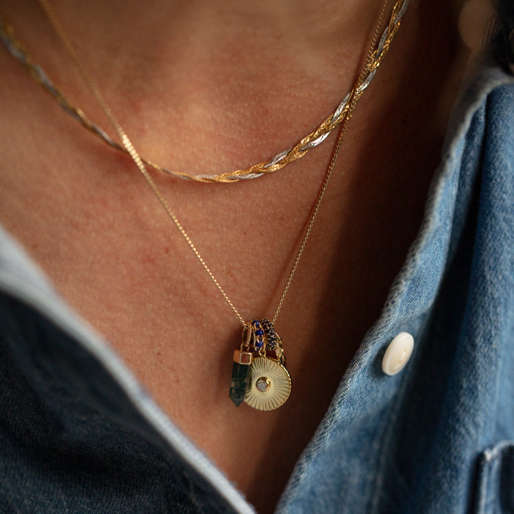 A close-up of a person wearing layered gold necklaces, including the Solid Gold Anniversaire Charm and other pendants, over a partially unbuttoned blue denim shirt.