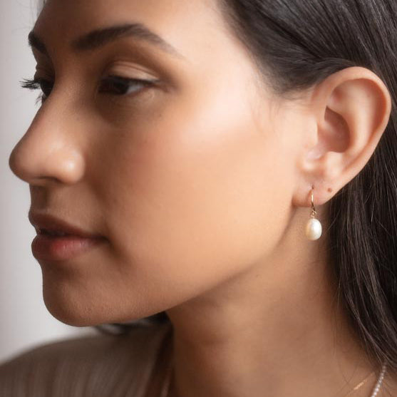 A close-up side view of a woman with smooth skin wearing the Solid Gold Elegant Pearl Charm Huggies on her left ear, featuring a removable pearl charm and her dark hair neatly tucked behind her ear.