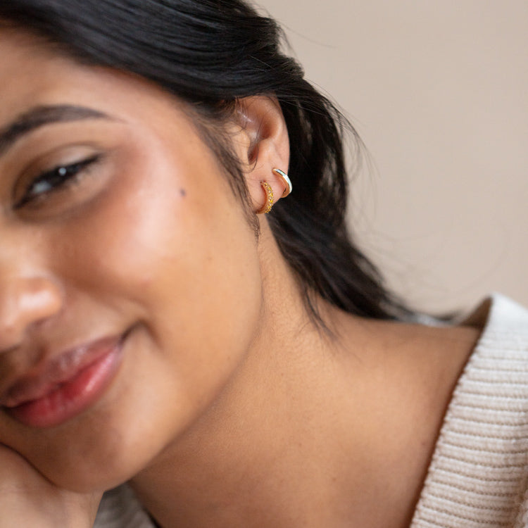 A woman with long dark hair smiles softly, resting her face on her hand. She wears Solid Gold Limited Edition Yellow Diamond Huggie Hoops and a cream-colored knit sweater, with a blurred neutral background.