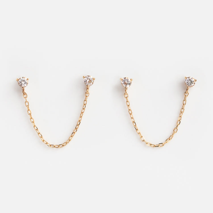 Solid Gold Double Piercing Chain Earrings, made from 9k solid gold, feature sparkling white topaz studs linked by a delicate gold chain. Displayed on a white background, each earring connects two dazzling studs with elegant style.