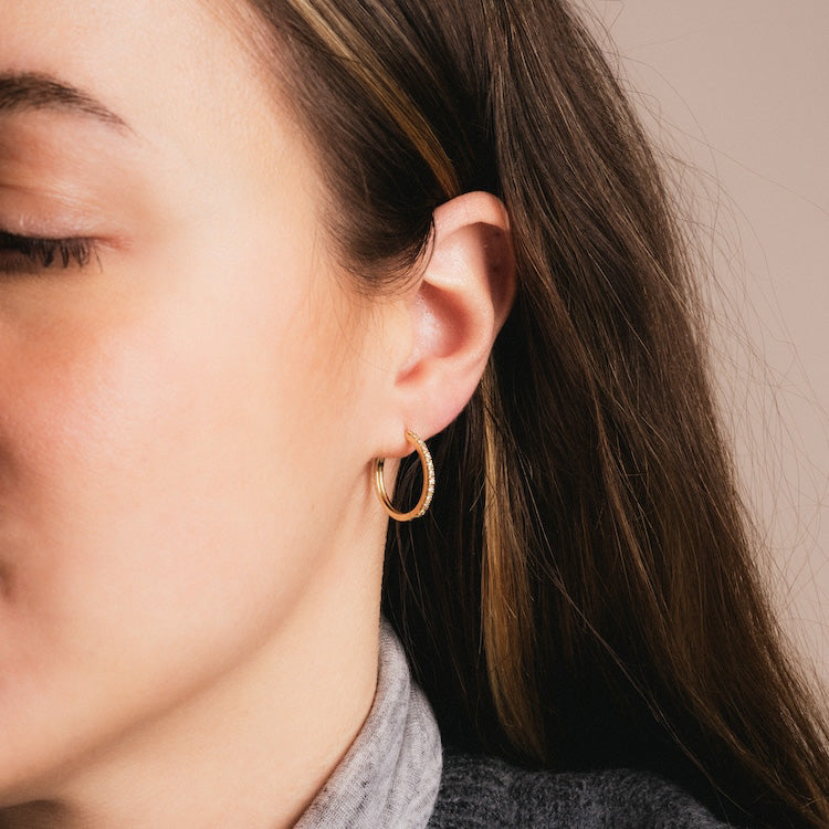 Close-up of a woman in profile with closed eyes, wearing Solid Gold Diamond Hoops. She has long brown hair and a gray collared shirt, set against a soft beige background.