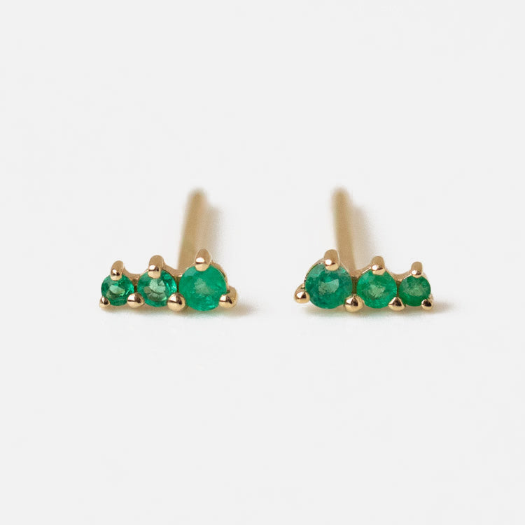 A pair of Solid Gold Dainty Birthstone Bar Studs, each showcasing three small round green gemstones in a row, displayed on a white background.