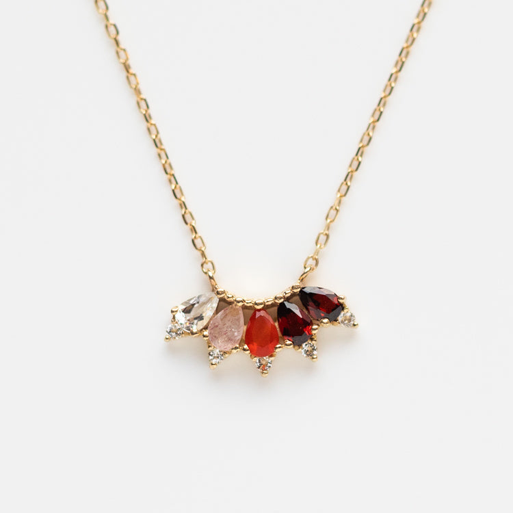 The Solid Gold Ombre Birthstone Necklace January Sample features a gold chain with five teardrop gemstones in clear, pink, red, and dark red—accented by natural garnet and small clear stones arranged in a fan shape.