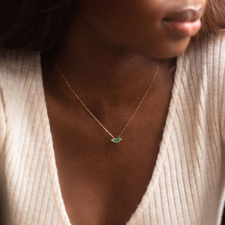 A woman in a cream, ribbed V-neck top showcases the Solid Gold Ombre Birthstone Necklace June Sample, a 9k yellow gold piece featuring natural alexandrite gemstones. The photo highlights her neckline and the delicate jewelry.