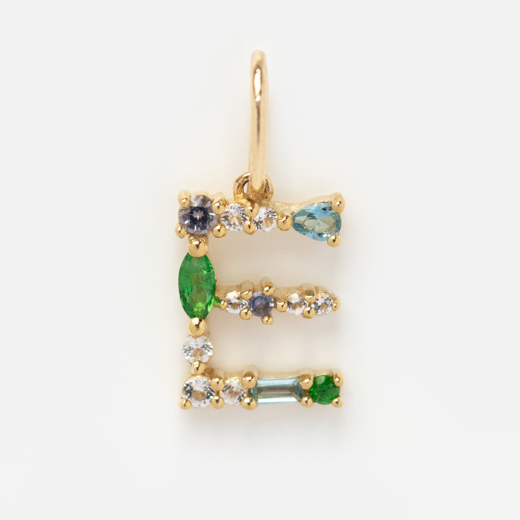 The Solid Gold Gemstone Letter Charm is crafted in solid gold and shaped like the letter E, featuring green, blue, and clear gemstones arranged along its elegant lines.