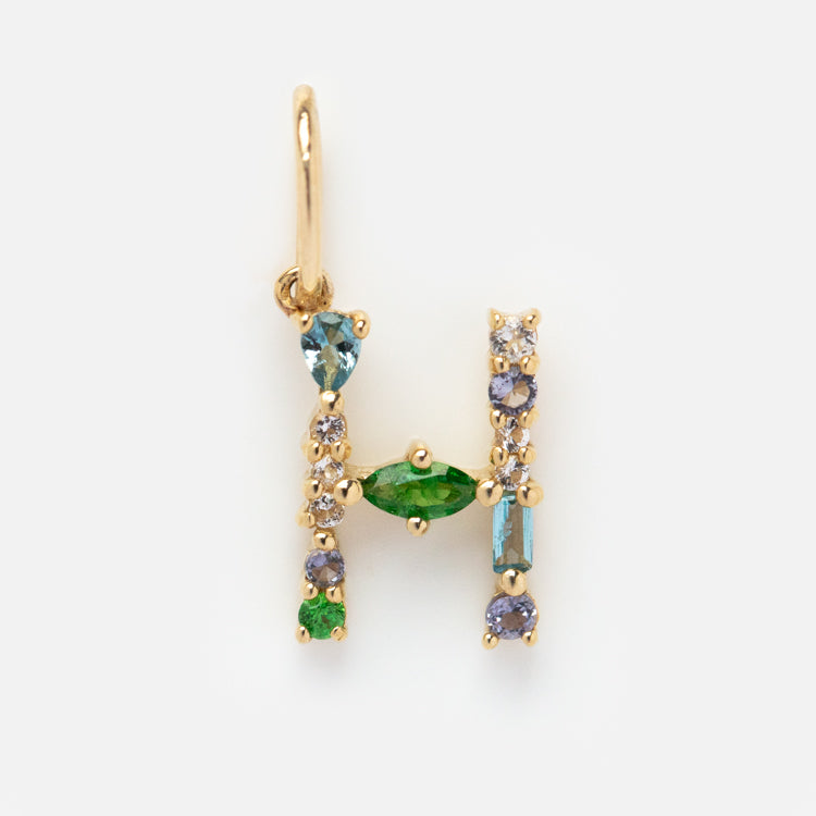 The Solid Gold Gemstone Letter Charm features an H-shaped pendant adorned with green, blue, and clear gemstones, displayed on a plain white background.