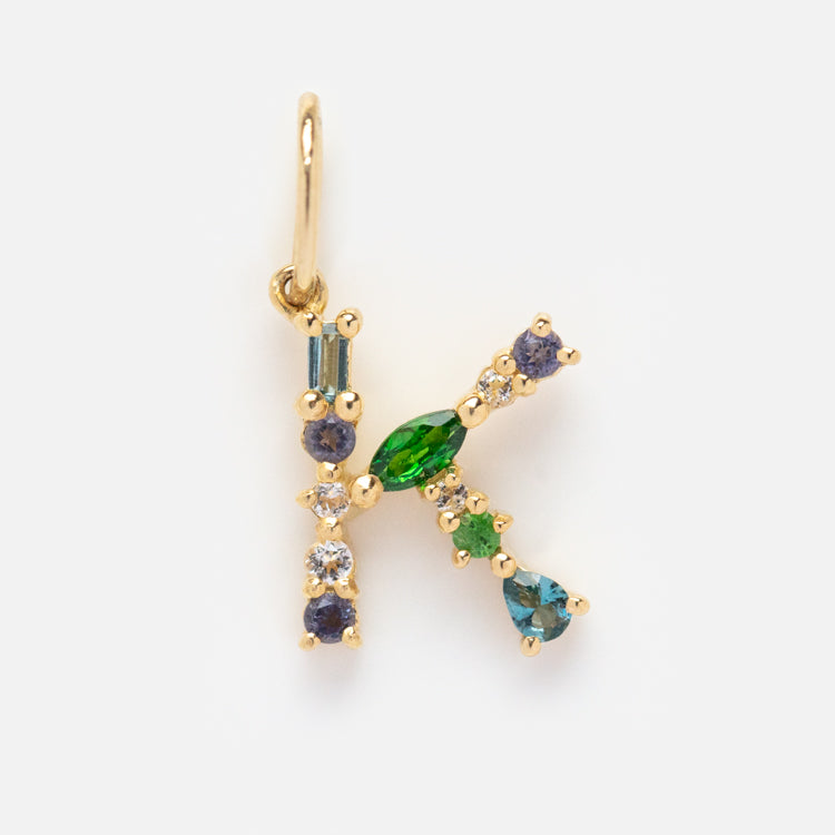 The Solid Gold Gemstone Letter Charm features the letter K, crafted in gold and embellished with green, blue, and clear gemstones, shown on a plain white background.