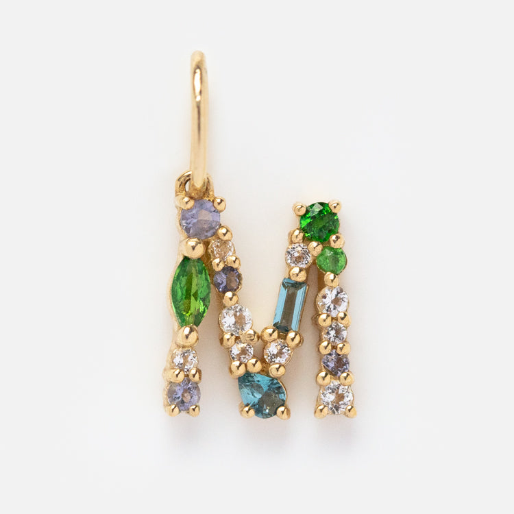 The Solid Gold Gemstone Letter Charm features the letter M, crafted in solid gold and adorned with green, blue, and clear gemstones, set against a plain white background.