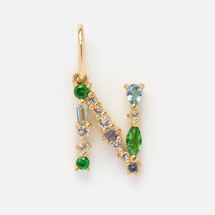 The Solid Gold Gemstone Letter Charm features the letter N in gold, embellished with green, blue, and clear gemstones of varying shapes and sizes, displayed on a plain white background.