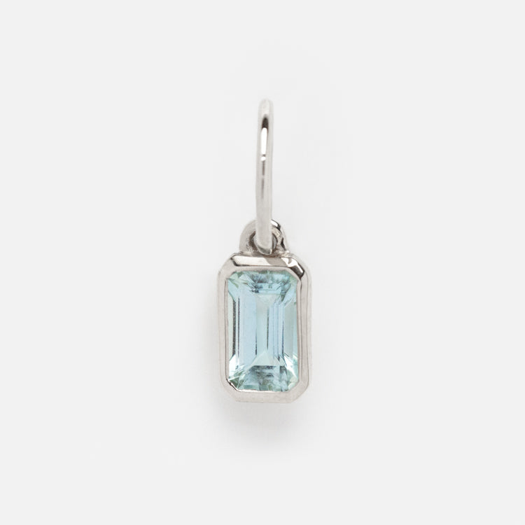 The Solid Gold Birthstone Baguette Charm in White Gold features a rectangular, emerald-cut light blue gemstone set in a sleek white gold frame with a small loop, perfect for personalizing your jewelry collection. Shown on a plain white background.