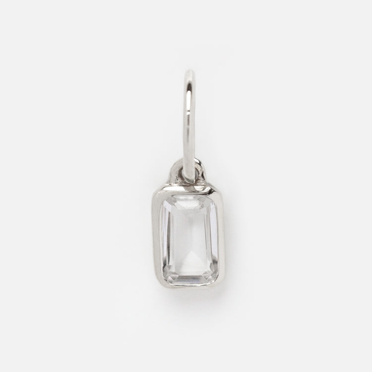 The Solid Gold Birthstone Baguette Charm in White Gold features a clear, faceted gemstone set at the center and a small loop for easy attachment, making it a stunning personalized jewelry addition. Shown on a plain white background.