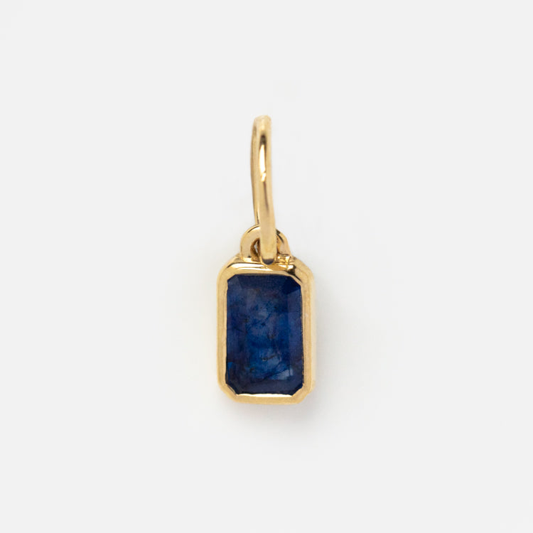 The Solid Gold Birthstone Baguette Charm features a small rectangular deep blue gemstone set in a simple gold bezel with a gold loop for attaching to necklaces or bracelets, shown against a plain white background.