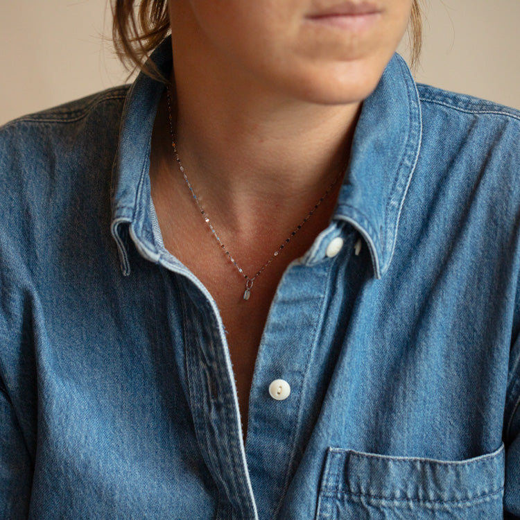 A person wearing a blue denim button-up shirt and a delicate silver necklace with the Solid Gold Birthstone Baguette Charm in White Gold. Shown from the shoulders up, their face is partially visible and turned slightly to the side.