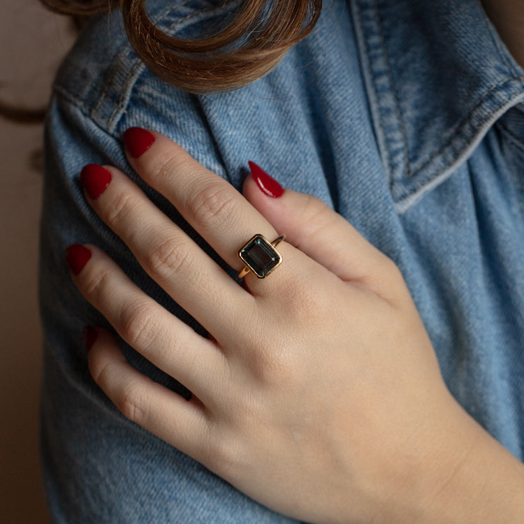 A hand with red-painted nails rests on a denim-clad arm, wearing the North South Bold Bezel Set Ring—a gold statement piece featuring a rectangular dark stone.
