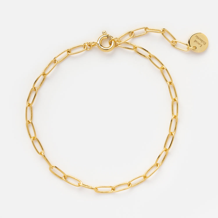 The Vermeil Paperclip Bracelet is a delicate 14k gold piece with elongated oval links, a round clasp, and an engraved tag, shown elegantly on a plain white background.